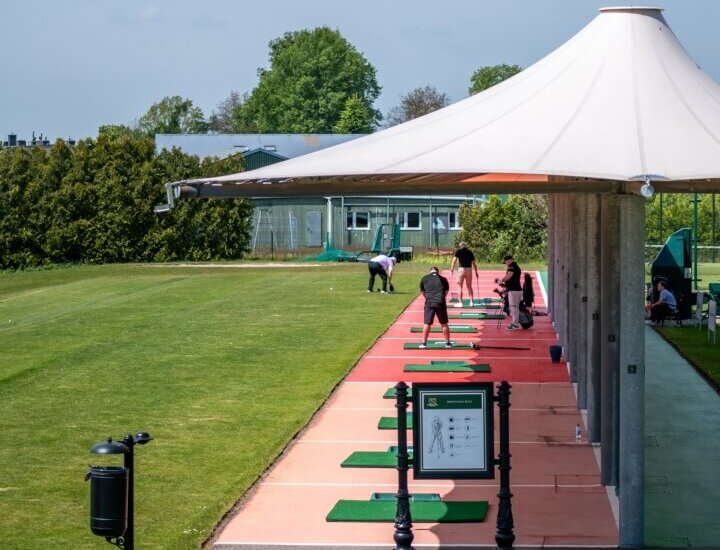 Zadaszona strzelnica golfowa z rzędami stanowisk treningowych na matach, goście ćwiczą uderzenia na zielonym polu