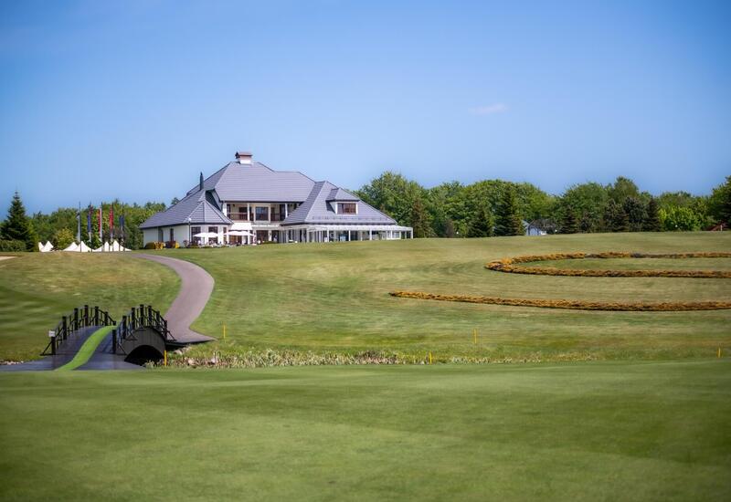 Klub golfowy z białym klubem i tarasem z parasolami, mostek nad wodą, szerokie zadbane trawniki i ozdobne okręgi kwiatowe.
