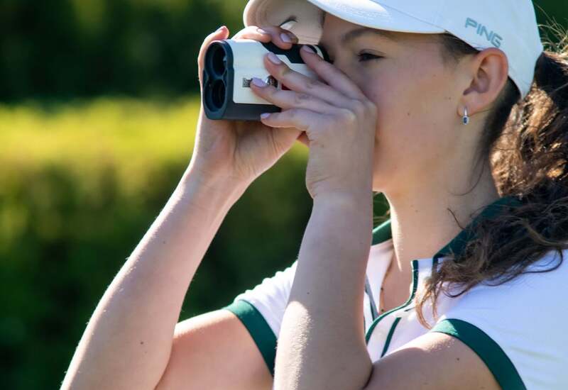 Golfistka na hotelowym polu golfowym mierzy odległość dalmierzem; biała czapka, sportowa koszulka i słoneczne, zielone tło