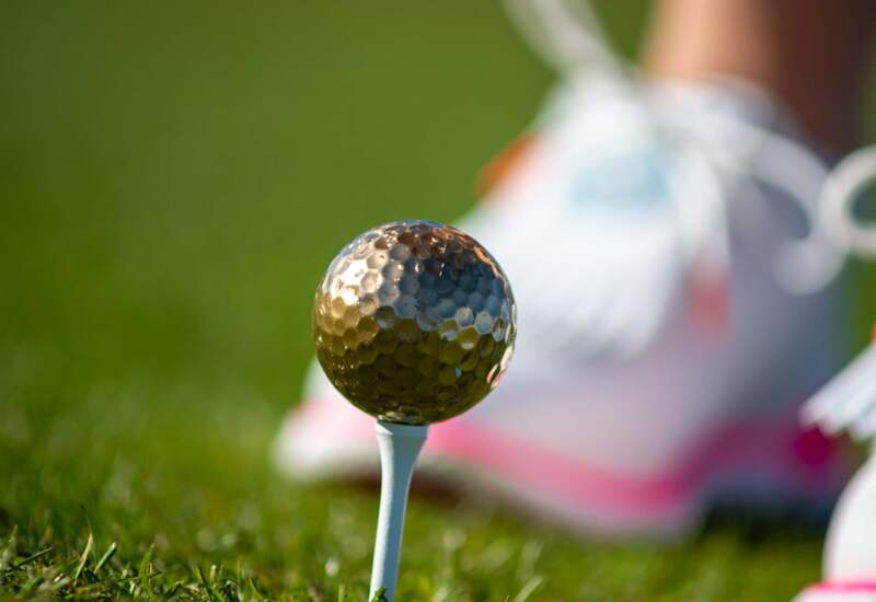 Złota piłka golfowa na tee na zielonym polu, dłoni kobiety ustawiającej ją przed uderzeniem, biało-różowe buty.
