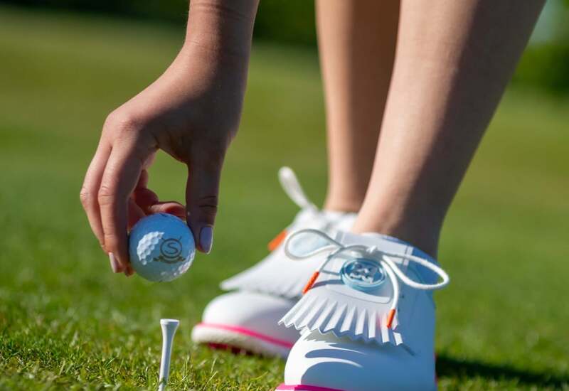 dłoń ustawiająca piłkę golfową na tee na zielonym polu, białe buty golfowe z różową podeszwą i ozdobnymi frędzlami