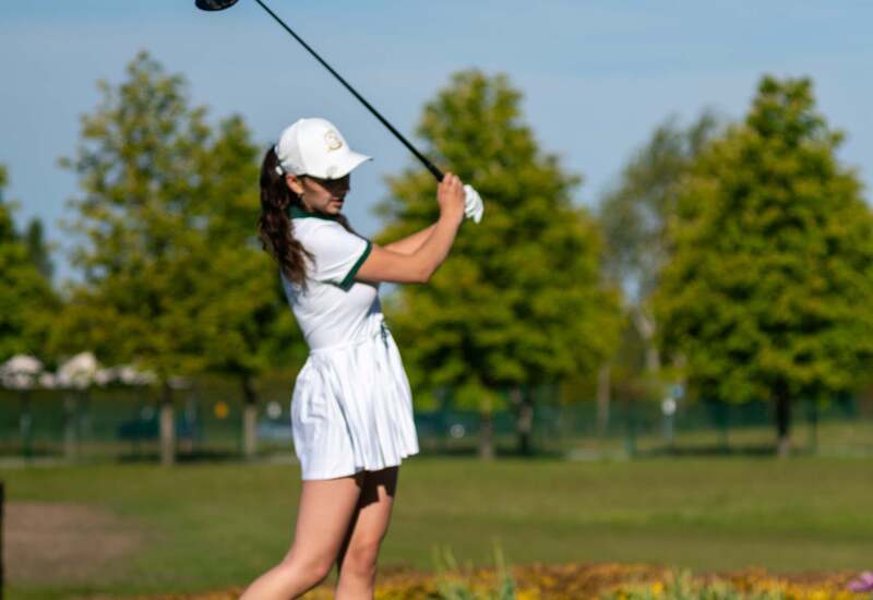 Kobieta uderza z tee na słonecznym polu golfowym kurortu — biała sportowa sukienka, zadbana trawa i drzewa w tle