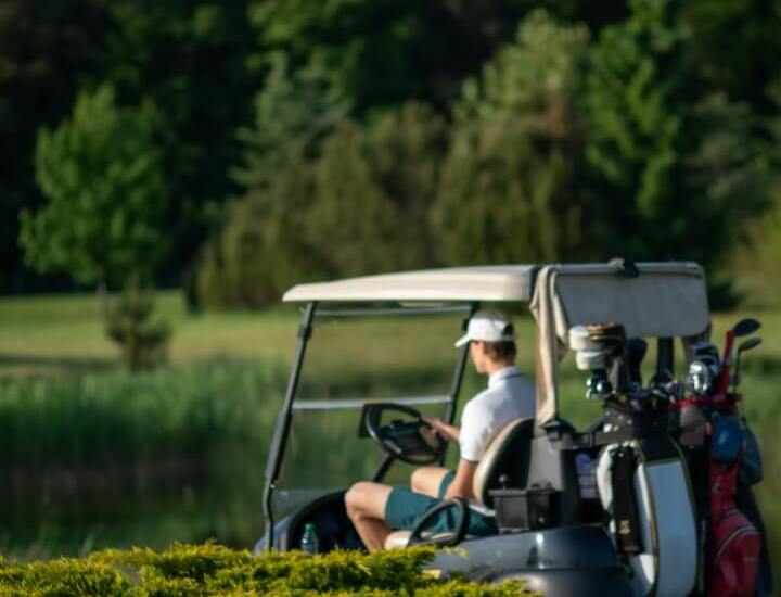 Gość na wózku golfowym z torbami i kijami na zielonym, jeziornym polu golfowym otoczonym lasem