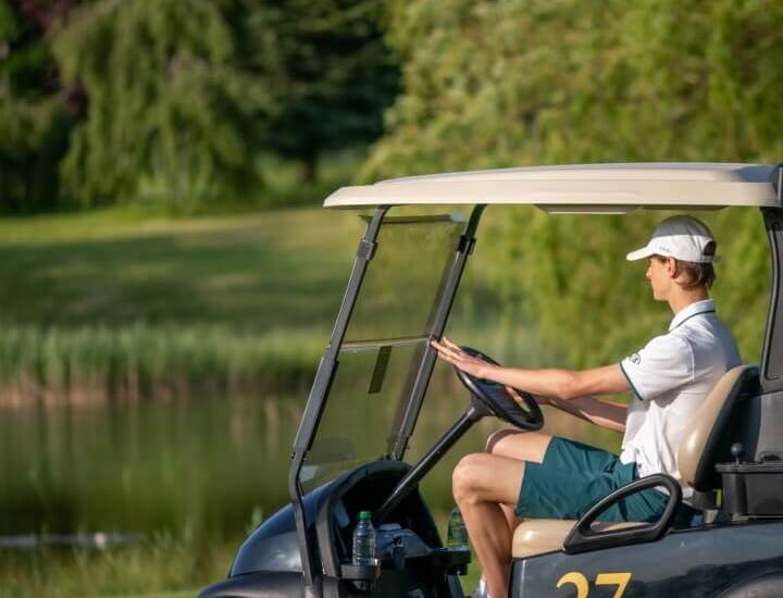 Gracz w koszulce polo siedzi w wózku golfowym nr 27 na zielonym polu przy stawie, spokojna, słoneczna atmosfera