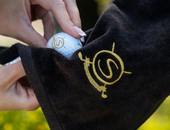 Ręce wkładają piłkę golfową do czarnego ręcznika z złotym monogramem hotelu, na tle zieleni pola golfowego