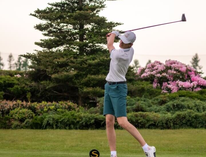 Golfista uderza z tee na hotelowym polu golfowym, zadbana trawa, sosna i kwitnące różowe rododendrony.