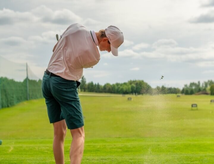 Golfista na polu treningowym uderza piłkę; trawa i kurz unoszą się, kosz z piłkami i szerokie zielone pole w tle