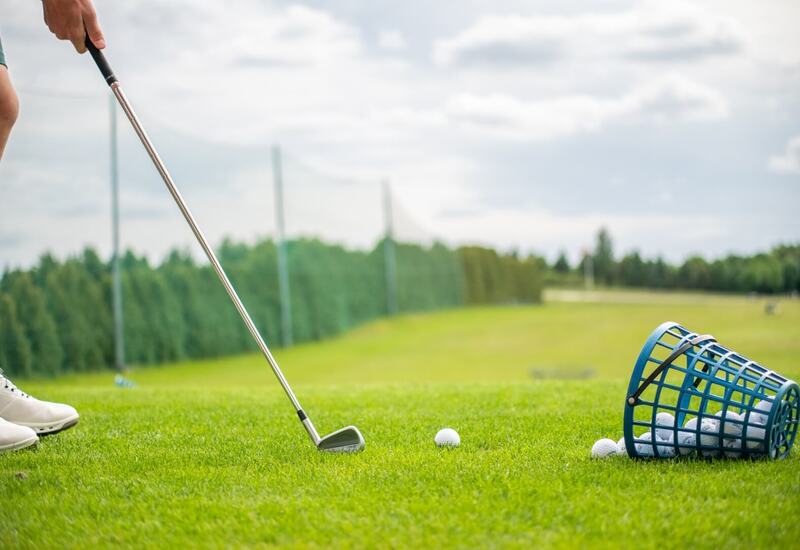 Golfista przygotowuje uderzenie na polu treningowym, przewrócony kosz z piłkami na zielonej trawie.