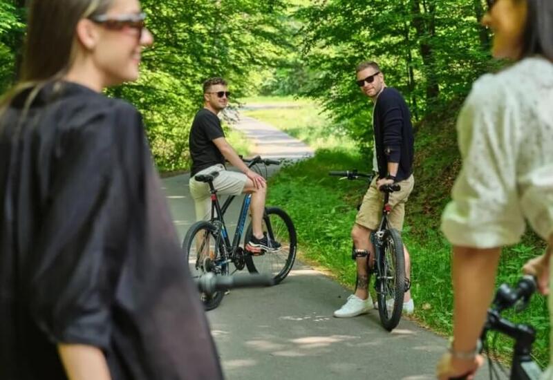 Cztery osoby na rowerach na leśnej ścieżce, dwoje mężczyzn odwraca się uśmiechając, letnia zacieniona trasa.