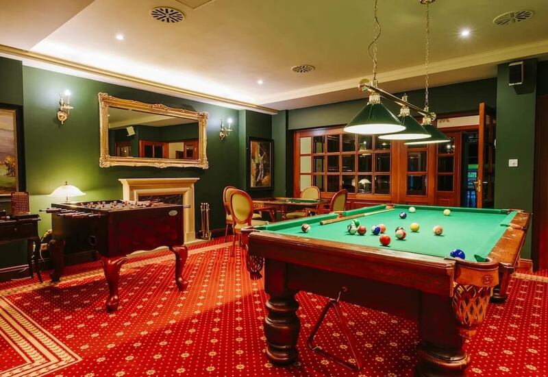 Sala gier hotelu z zielonym stołem bilardowym, czerwonym dywanem, wiszącymi lampami, piłkarzykami i kominkiem.
