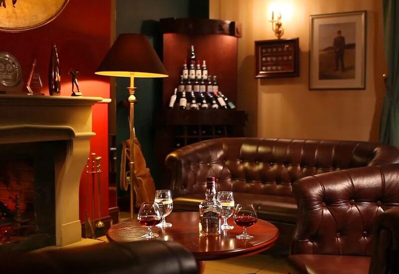 Przytulny salon hotelowy z kominkiem, skórzanymi kanapami Chesterfield, stolikiem z butelką whisky i kieliszkami oraz regałem win.