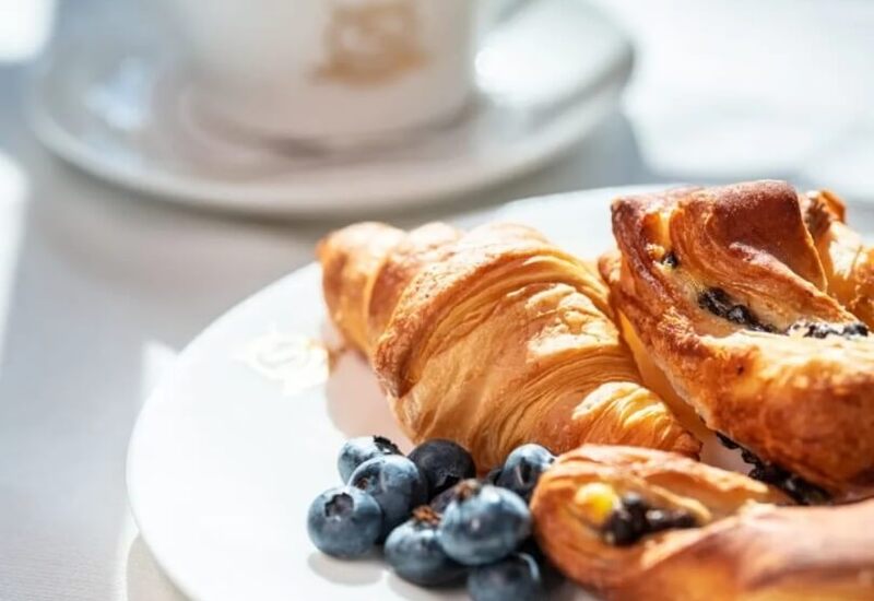 Świeże croissanty i rogaliki z borówkami na białym talerzu, obok filiżanka cappuccino w miękkim porannym świetle