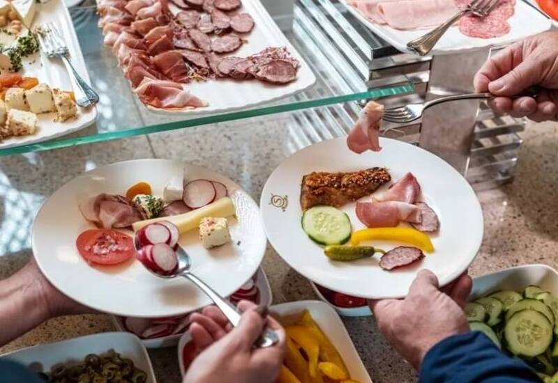 Bufet śniadaniowy z wędlinami i serami, płatkami, misami z papryką, ogórkami i oliwkami; goście nakładają jedzenie.
