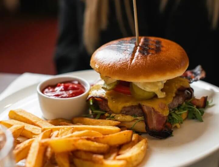 Burger w bułce brioche z topionym serem, bekonem i ogórkami, podany z chrupiącymi frytkami i ketchupem na białym talerzu w restauracji