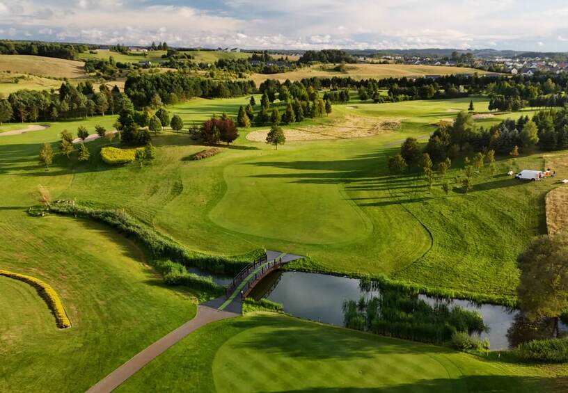 Widok pola golfowego z zielonymi dołkami, bunkrami, drewnianym mostkiem nad stawem i rozległymi wiejskimi widokami
