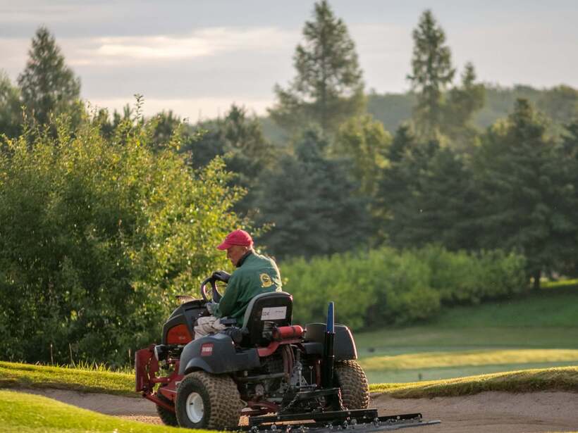 Pracownik obsługi trawników na traktorku pielęgnacyjnym wygładza bunkier na zielonym polu golfowym, poranne światło.