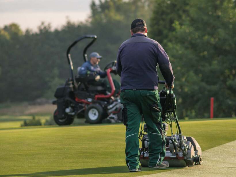 Pracownik pielęgnuje idealnie przystrzyżoną zieloną nawierzchnię pola golfowego maszyną do koszenia, pojazd serwisowy w tle.
