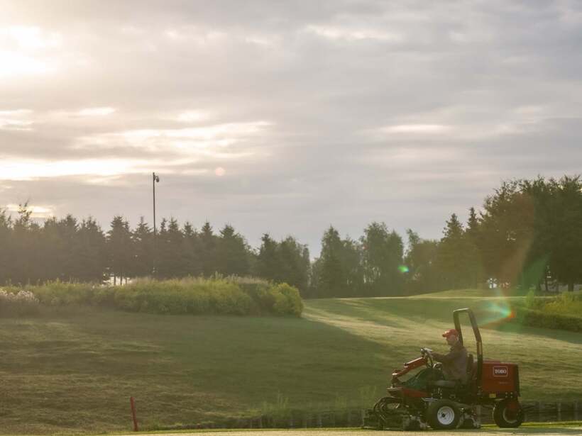 Wschód nad zadbanym polem golfowym, pracownik kosi trawę traktorkiem, równo przycięte fairwaye i rosa.