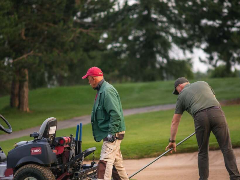 Pracownicy pola golfowego wyrównują bunkier obok pojazdu terenowego, zadbana trawa i otaczające drzewa.