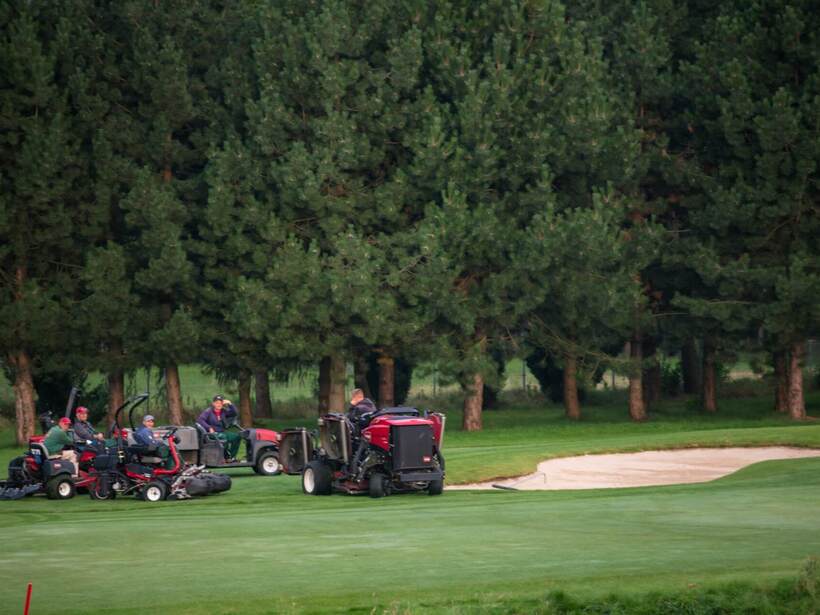Ekipa utrzymania pola golfowego z kosiarkami i traktorkami przy zielonej trawie i piaskowym bunkrze pod wysokimi drzewami.