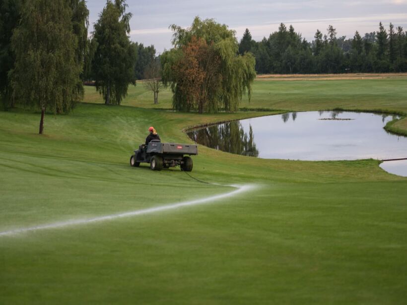 Personel na wózku serwisowym podlewa zadbane pole golfowe przy stawie; równo skoszone trawniki i otaczające drzewa