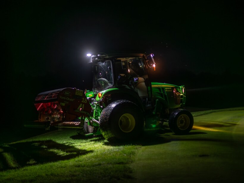 Traktor serwisowy na polu golfowym nocą, oświetlony reflektorami podczas aeracji trawnika.