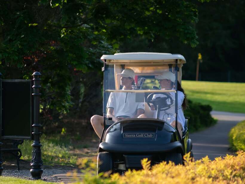 Para gości w wózku golfowym Sierra Golf Club jedzie ścieżką między zielonymi drzewami.
