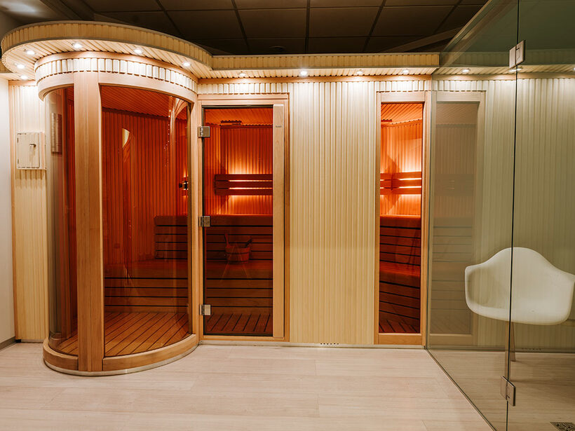 Hotelowa sauna z drewnianą kabiną i szklanym zaokrąglonym wejściem, ciepłe oświetlenie, ławki i przeszklona strefa relaksu.