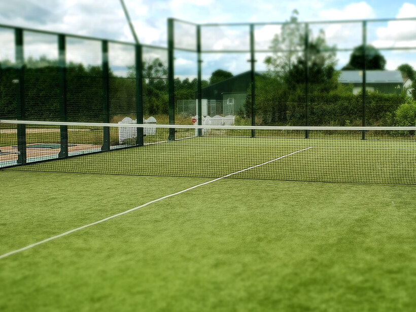 Kort padel na sztucznej trawie z siatką i szklanymi ściankami, z widokiem na zielone otoczenie i leżaki