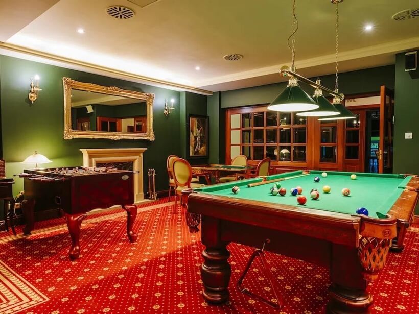 Sala gier hotelu z zielonym stołem bilardowym, czerwonym dywanem, wiszącymi lampami, piłkarzykami i kominkiem.