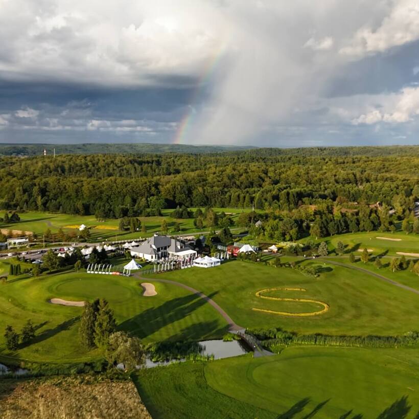 Rozległe pole golfowe z klubem, namiotami imprezowymi, stawami i mostkami, otoczone lasem, z tęczą nad horyzontem.