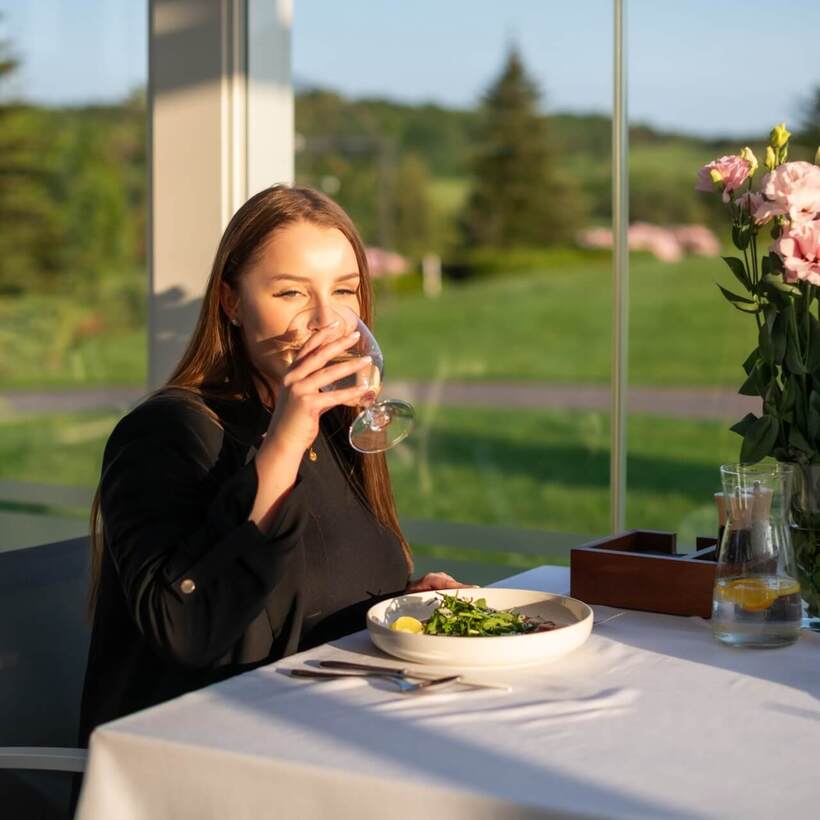 Kobieta pije wino w przeszklonej restauracji z talerzem sałatki, karafką z cytryną, bukietem różowych kwiatów i widokiem na ogród.