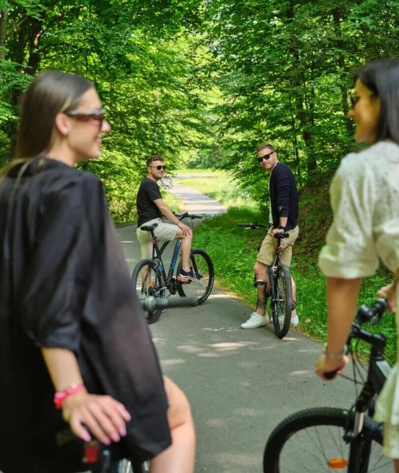 Cztery osoby na rowerach na leśnej ścieżce, dwoje mężczyzn odwraca się uśmiechając, letnia zacieniona trasa.