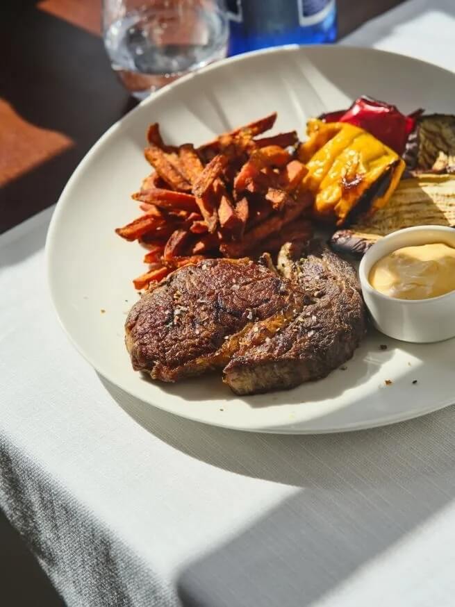 Stek z chrupiącą skórką, bataty w słupkach, grillowane warzywa i dip na białym talerzu, podane w słonecznym hotelowym bistro.