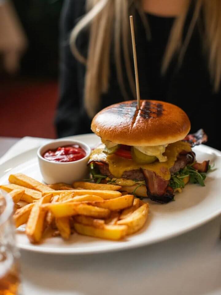 Burger w bułce brioche z topionym serem, bekonem i ogórkami, podany z chrupiącymi frytkami i ketchupem na białym talerzu w restauracji