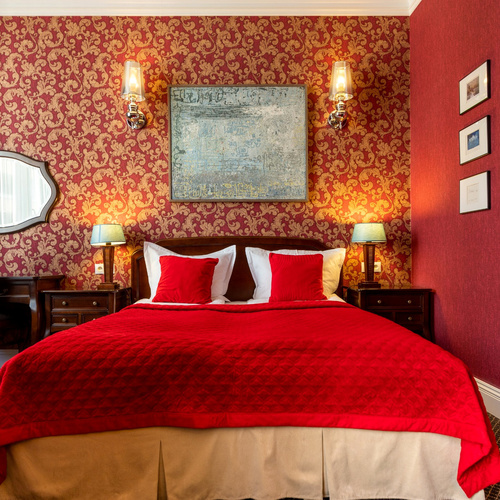 Front view of the double bed with a red bedspread, bedside lamps and patterned wallpaper in Junior Suite Deluxe Red