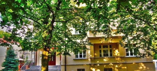 Wejście do hotelu pod rozłożystym drzewem, kamienne schody z poręczami, kremowa elewacja z balkonem i czerwonymi drzwiami