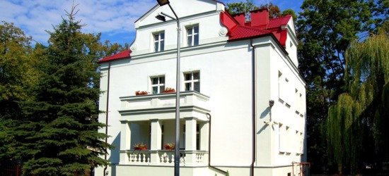 Biała willa z czerwonym dachem i balkonem z kwiatami, okrągły brukowany dziedziniec z ławkami i rabatą białych kwiatów