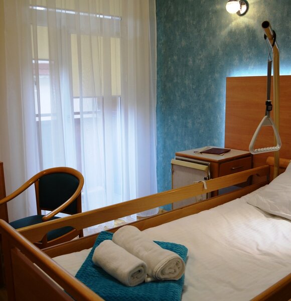 Pokój hotelowy przystosowany: regulowane łóżko z barierkami i uchwytem do podciągania, zrolowane ręczniki na niebieskiej narzucie, fotel, szafka, firanki