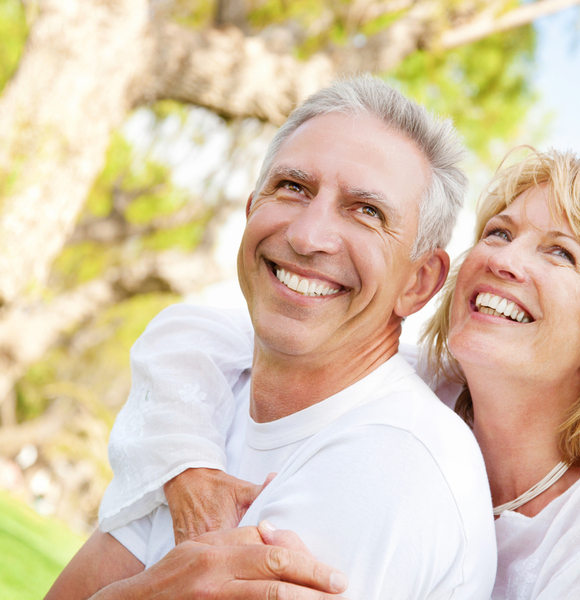 Lächelndes reifes Paar in weißen Hemden, Frau umarmt Mann von hinten im sonnigen Park unter einem großen Baum