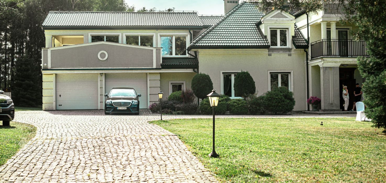 Luksusowa willa z eleganckim podjazdem, garażem, zielonym ogrodem i eleganckim wejściem z balkonem i latarniami.