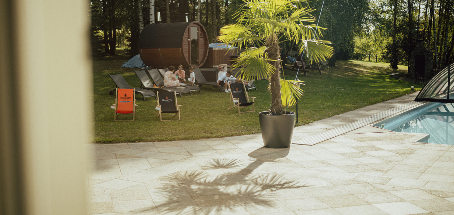 Strefa wypoczynkowa z leżakami, beczkową sauną i palmą doniczkową przy basenie na tle leśnej scenerii
