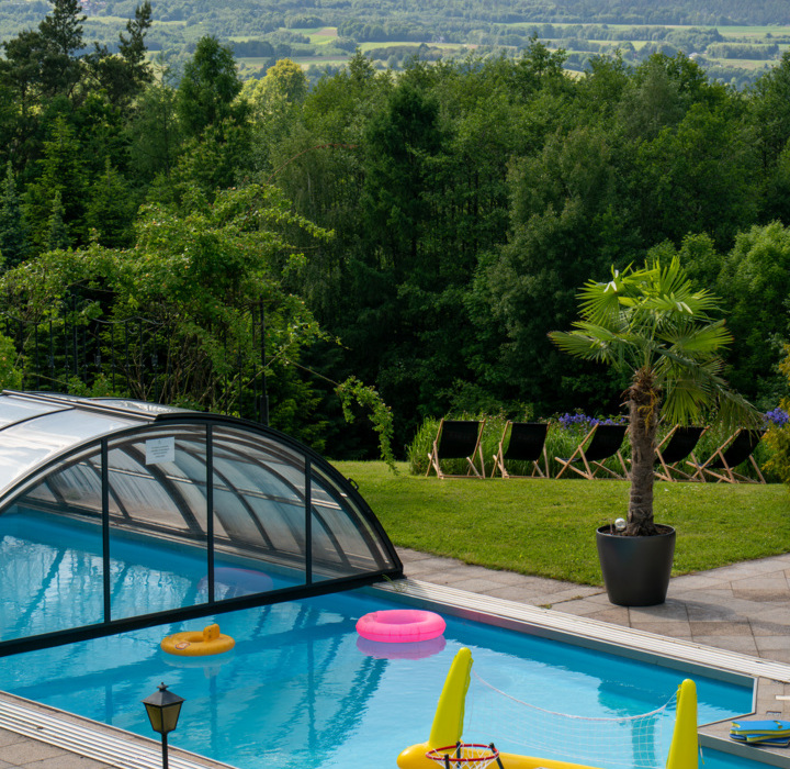 Basen z przezroczystym zadaszeniem, leżaki na trawie i palma doniczkowa na wypoczynkowym tarasie z widokiem na zalesione wzgórza.