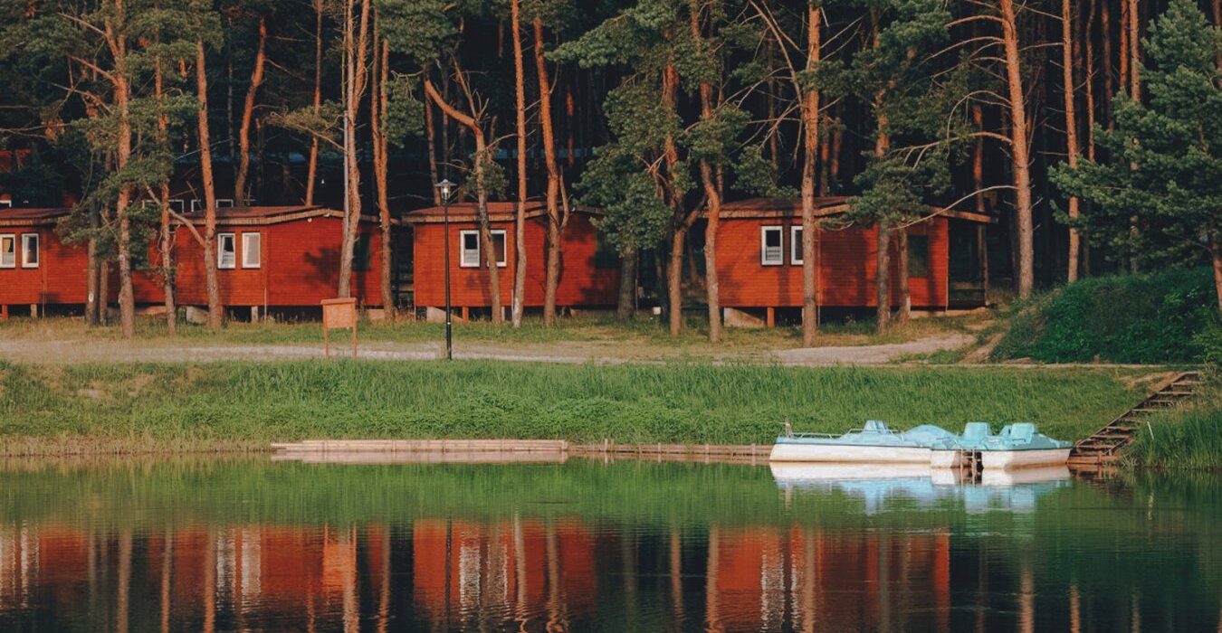 Drewniane domki letniskowe nad stawem z rowerkami wodnymi, otoczone sosnowym lasem.