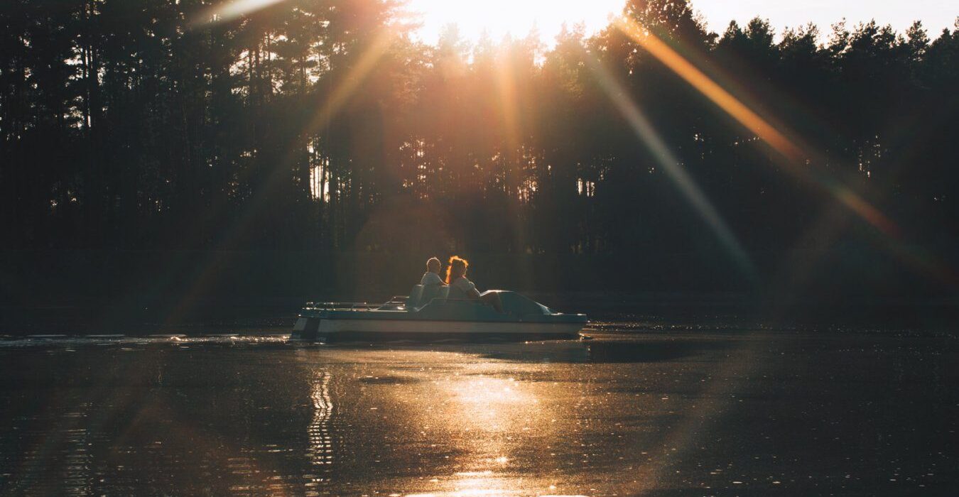 Dwoje gości na rowerku wodnym pedałujących po stawie o zachodzie słońca, otoczonym sosnowym lasem.