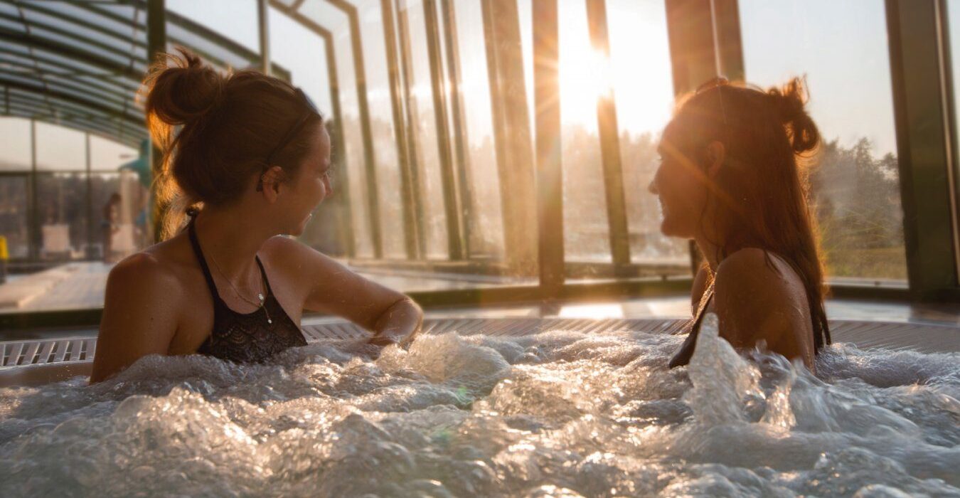 Dwie kobiety relaksujące się w jacuzzi w spa z widokiem na las i rzekę Wartę.