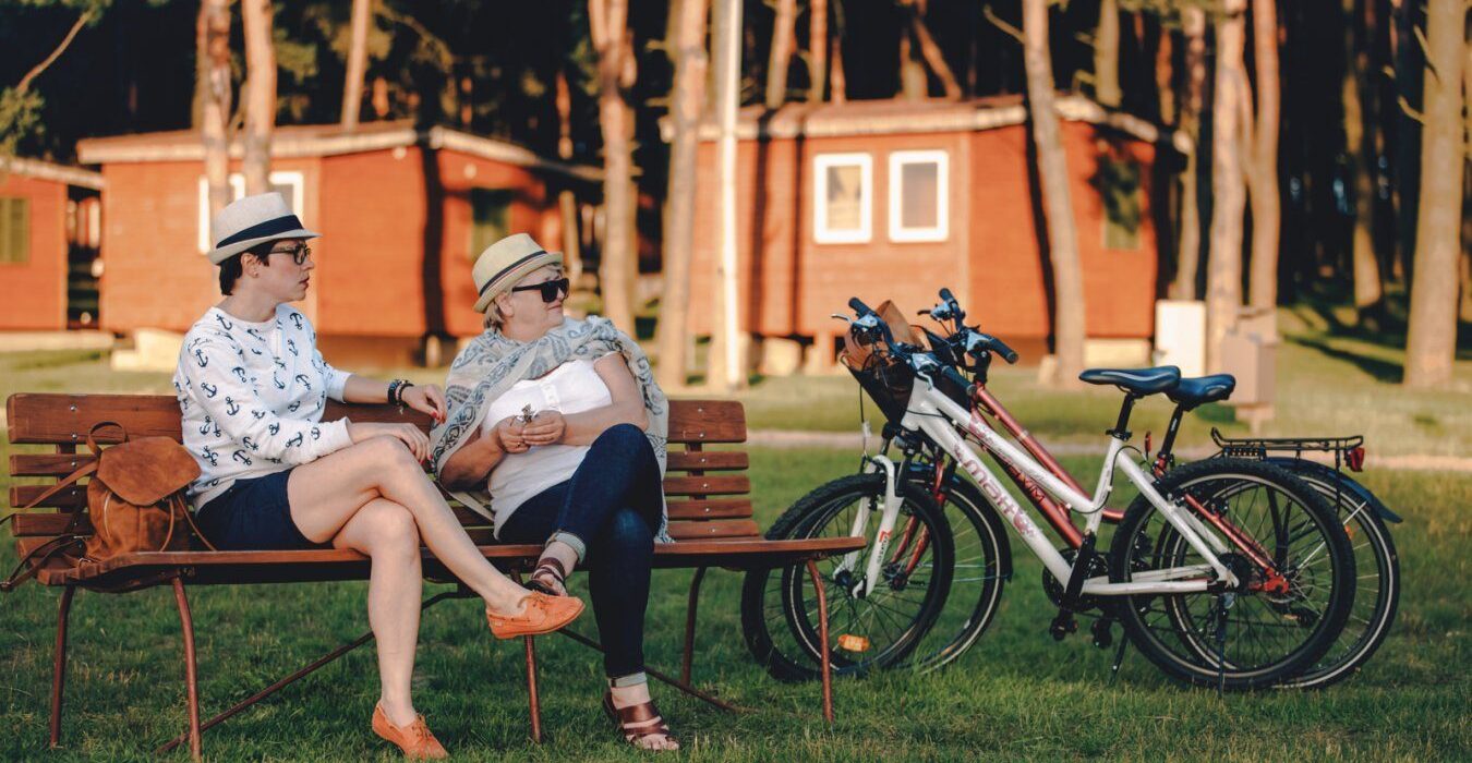 Dwie kobiety odpoczywające na ławce w Resort Stara Wieś przy drewnianych domkach letniskowych, obok stoją rowery.