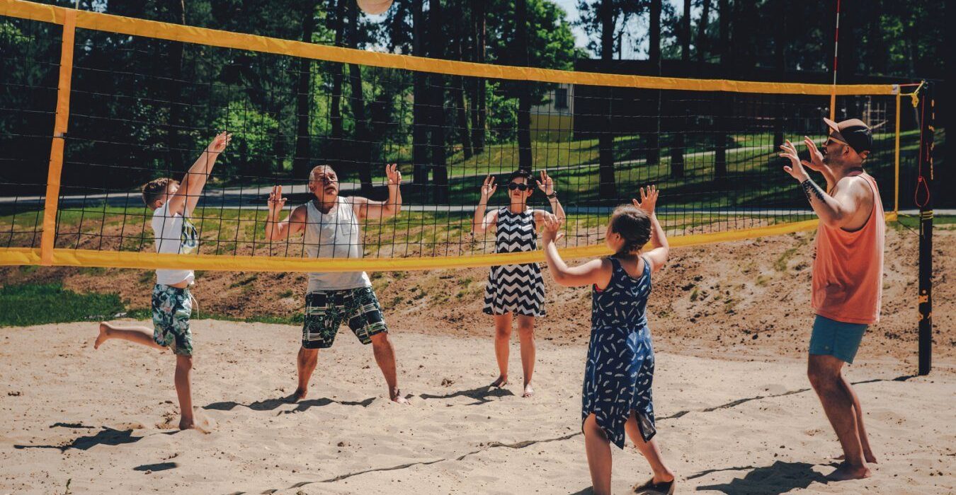 Rodzina grająca w siatkówkę plażową na piaszczystym boisku otoczonym zielonymi drzewami w słoneczny dzień.