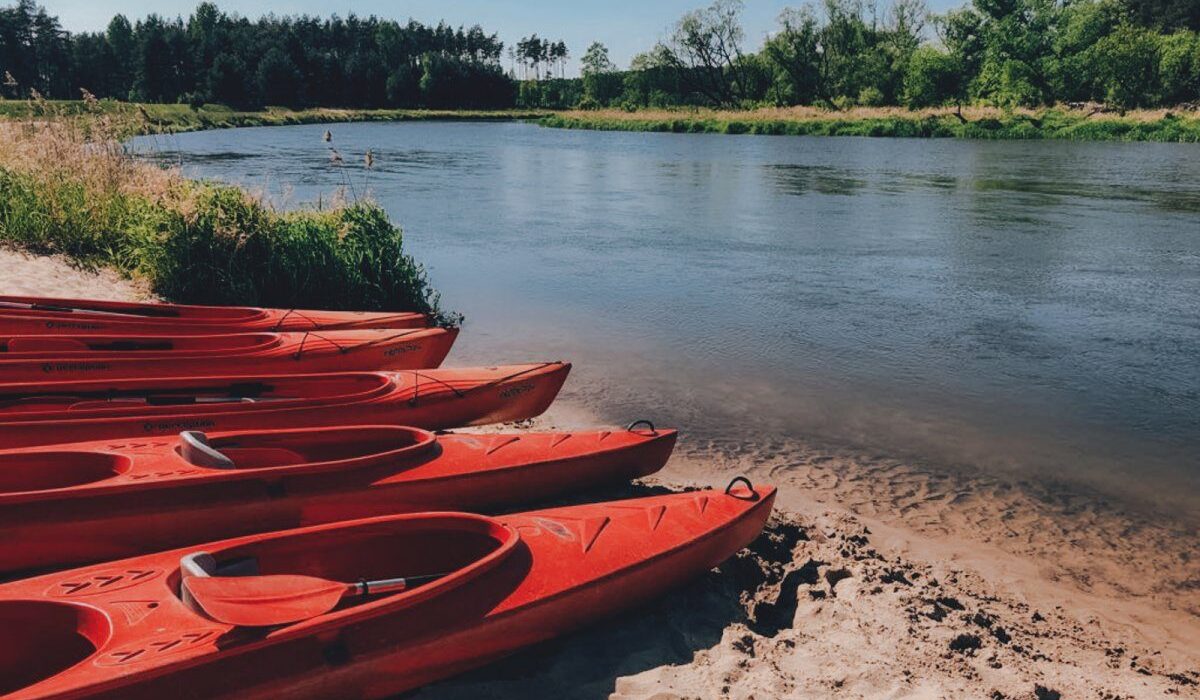 Czerwone kajaki ustawione na piaszczystej plaży nad spokojną rzeką Wartą otoczoną zielonymi drzewami.
