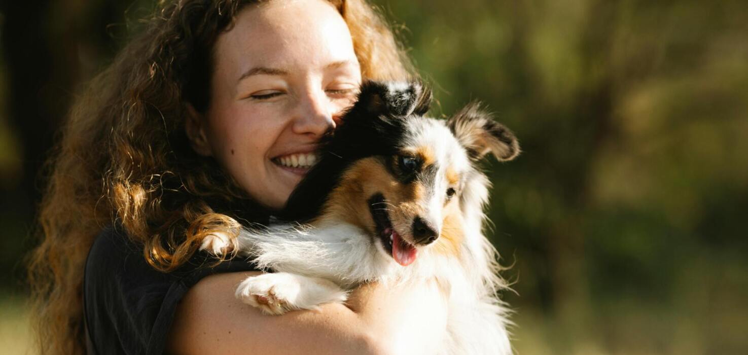 Uśmiechnięta kobieta z długimi kręconymi włosami przytulająca szczęśliwego psa na zewnątrz w słoneczny dzień.